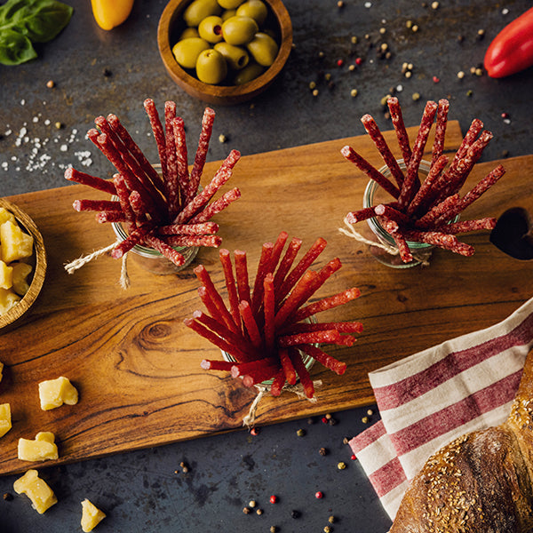Serviervorschlag Malbuner Party Sticks Poulet in 3 Gläsern auf Holzbrett mit Oliven, Paprika, Käse, Brot und Pfefferkörner