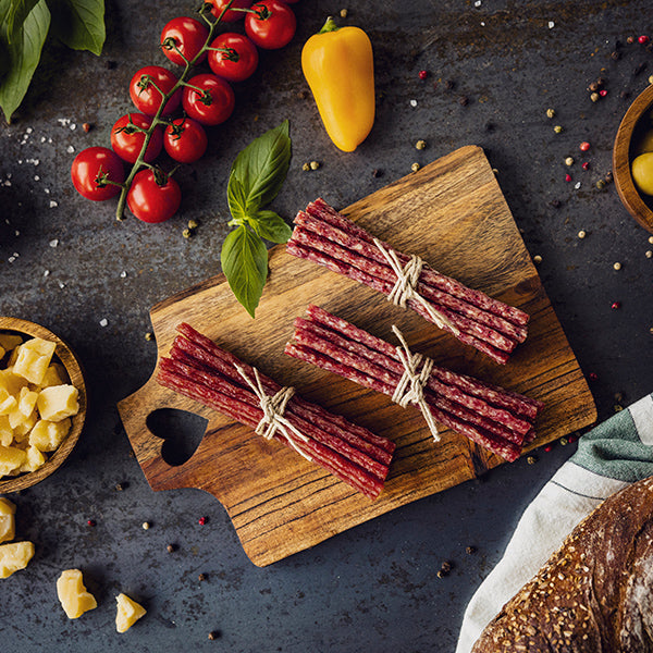 Serviervorschlag Malbuner Party Sticks Classic auf Holzbrett mit Tomaten, Paprika, Brot, Käse und Pfefferkörner