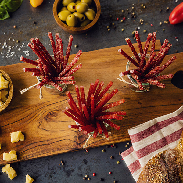 Serviervorschlag Malbuner Party Sticks Classic in 3 Gläsern auf Holzbrett mit Oliven, Paprika, Käse, Brot und Pfefferkörner